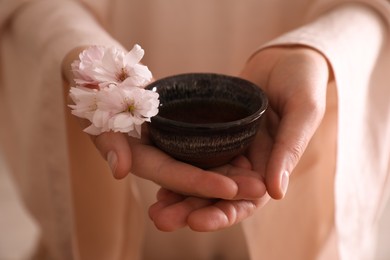 Photo of Master holding cup of freshly brewed tea and sakura flowers during traditional ceremony at table indoors, closeup
