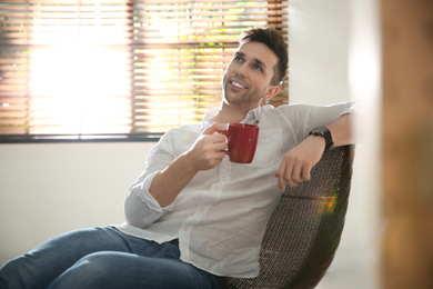 Photo of Young man with cup of drink relaxing near window at home