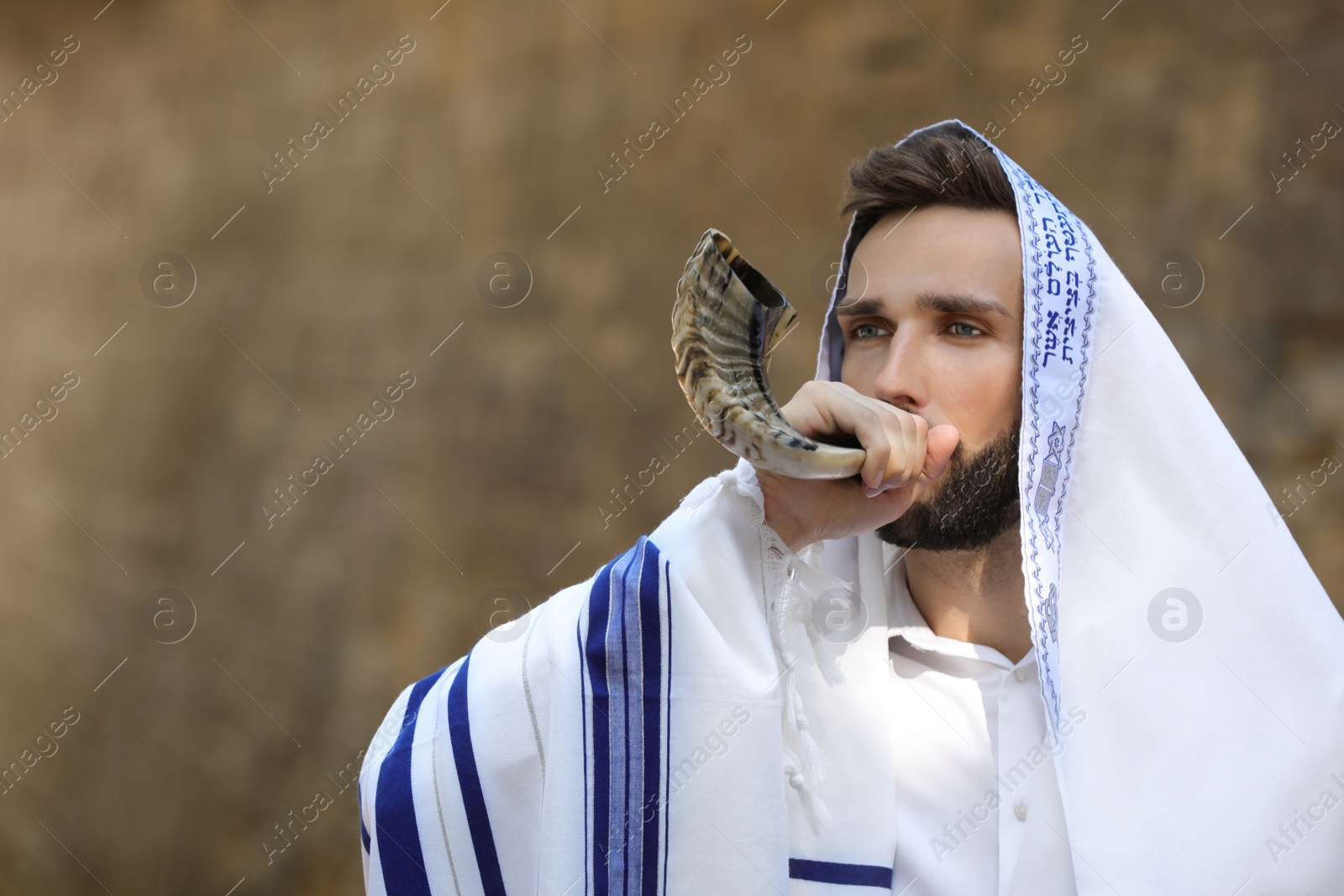 Photo of Jewish man blowing shofar on Rosh Hashanah outdoors. Wearing tallit with words Blessed Are You, Lord Our God, King Of The Universe, Who Has Sanctified Us With His Commandments, And Commanded Us To Enwrap Ourselves In Tzitziton in Hebrew