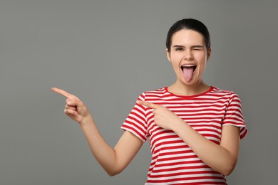 Photo of Happy young woman showing her tongue and pointing on grey background. Space for text