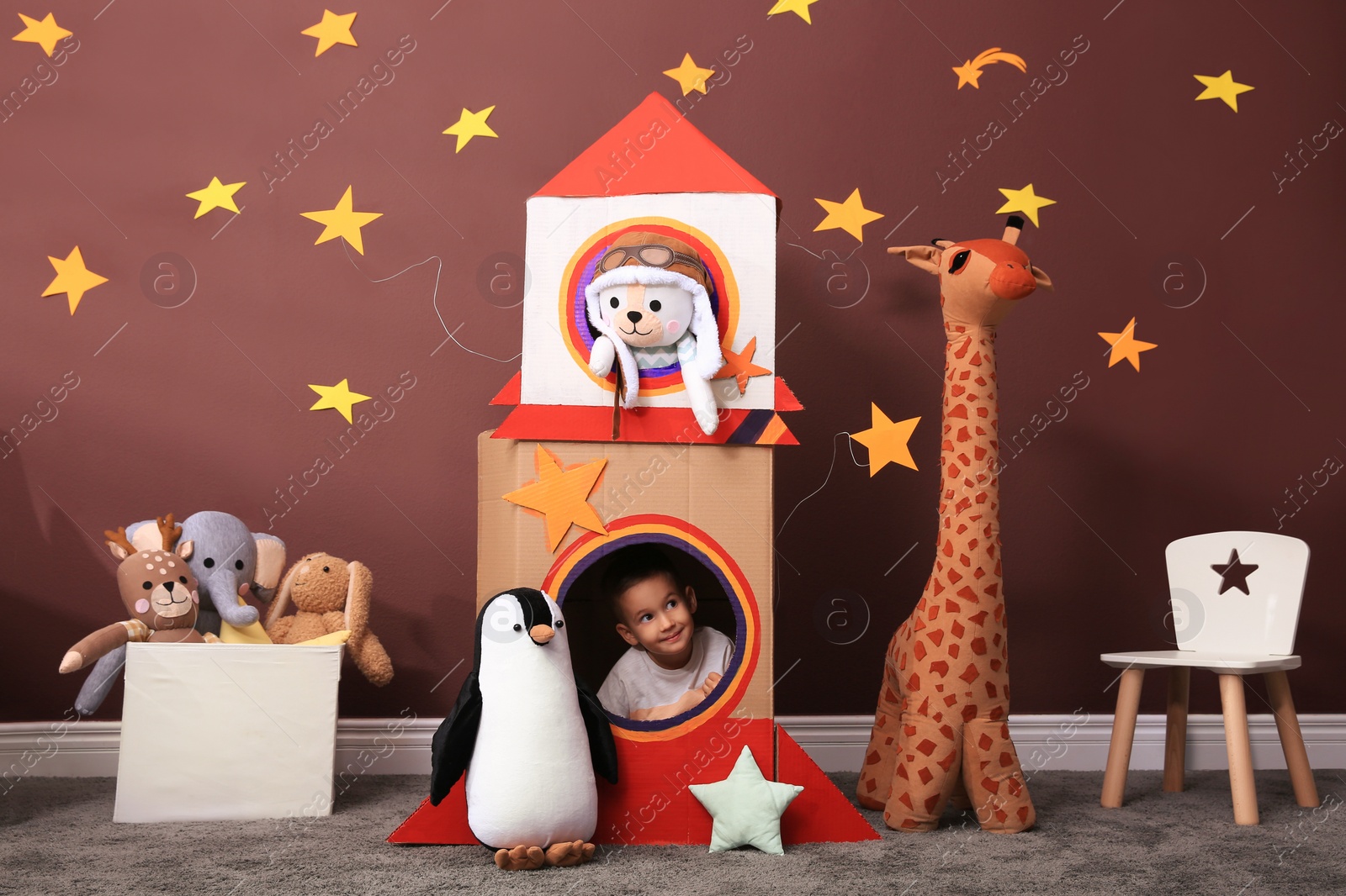 Photo of Cute little boy playing with cardboard rocket and toys at home. Child's room interior