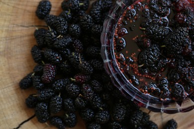 Delicious ripe black mulberries and bowl of sweet jam on wood stump, flat lay