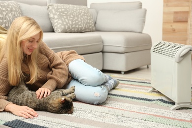 Young woman and cute tabby cat near electric heater at home