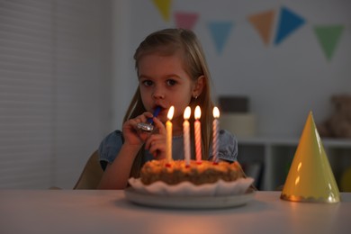 Photo of Birthday celebration. Cute girl holding blower at table with tasty cake indoors