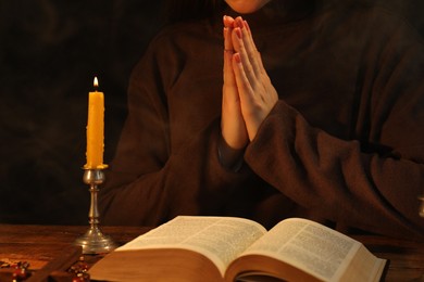 Woman praying at table with burning candle and Bible, closeup