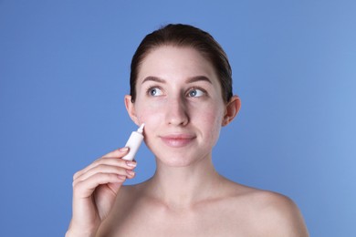 Young woman with acne problem applying cosmetic product onto her skin on blue background