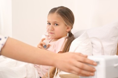 Mother helping her sick daughter with nebulizer inhalation in bedroom, closeup