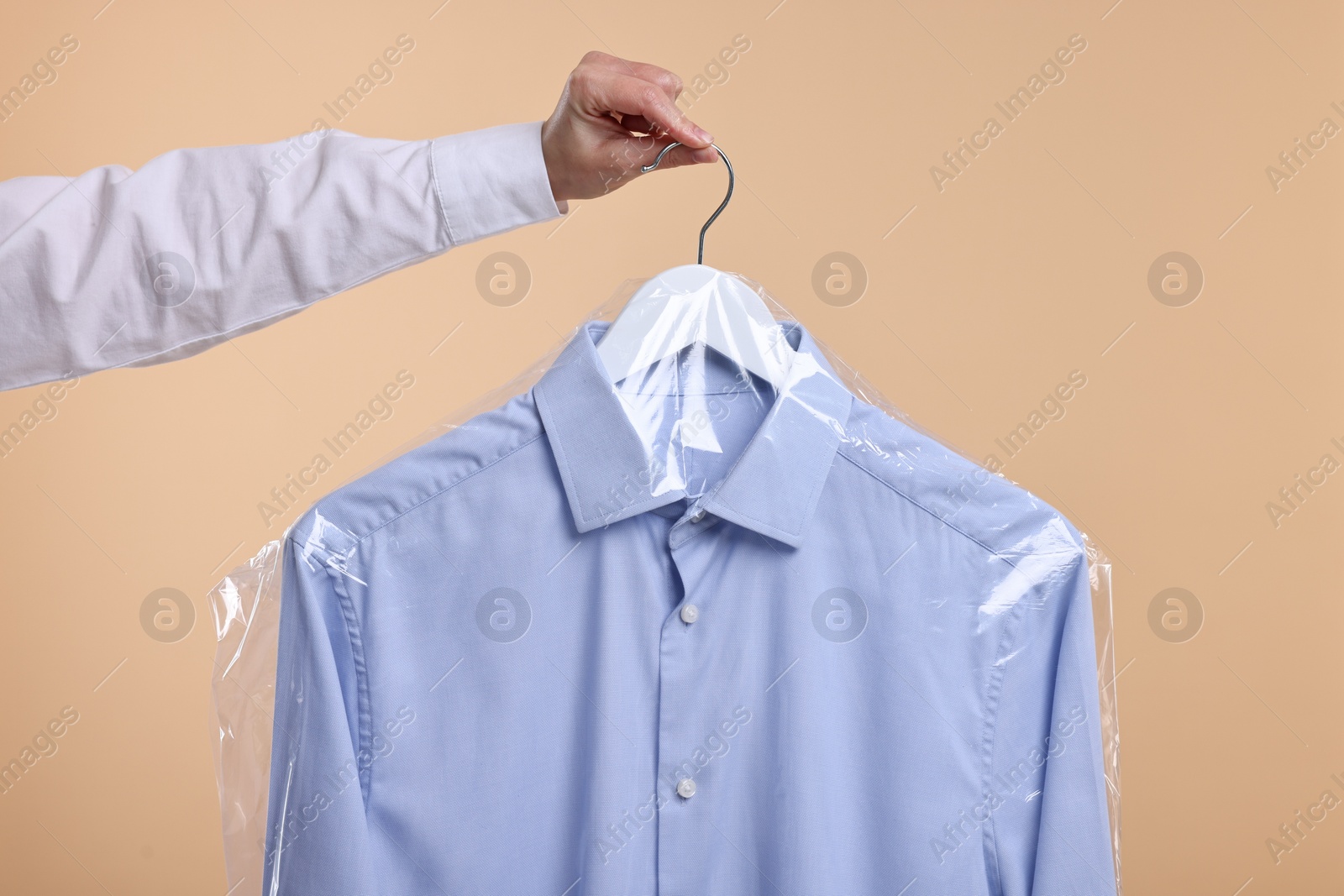 Photo of Dry-cleaning service. Woman holding shirt in plastic bag on beige background, closeup