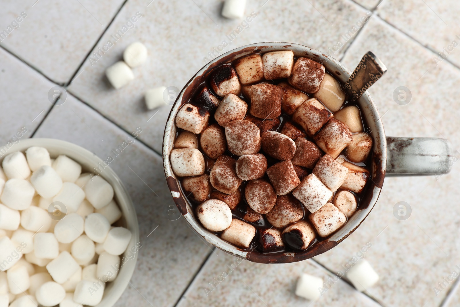 Photo of Delicious hot chocolate with marshmallows, cocoa powder and spoon in cup on tiled table, top view