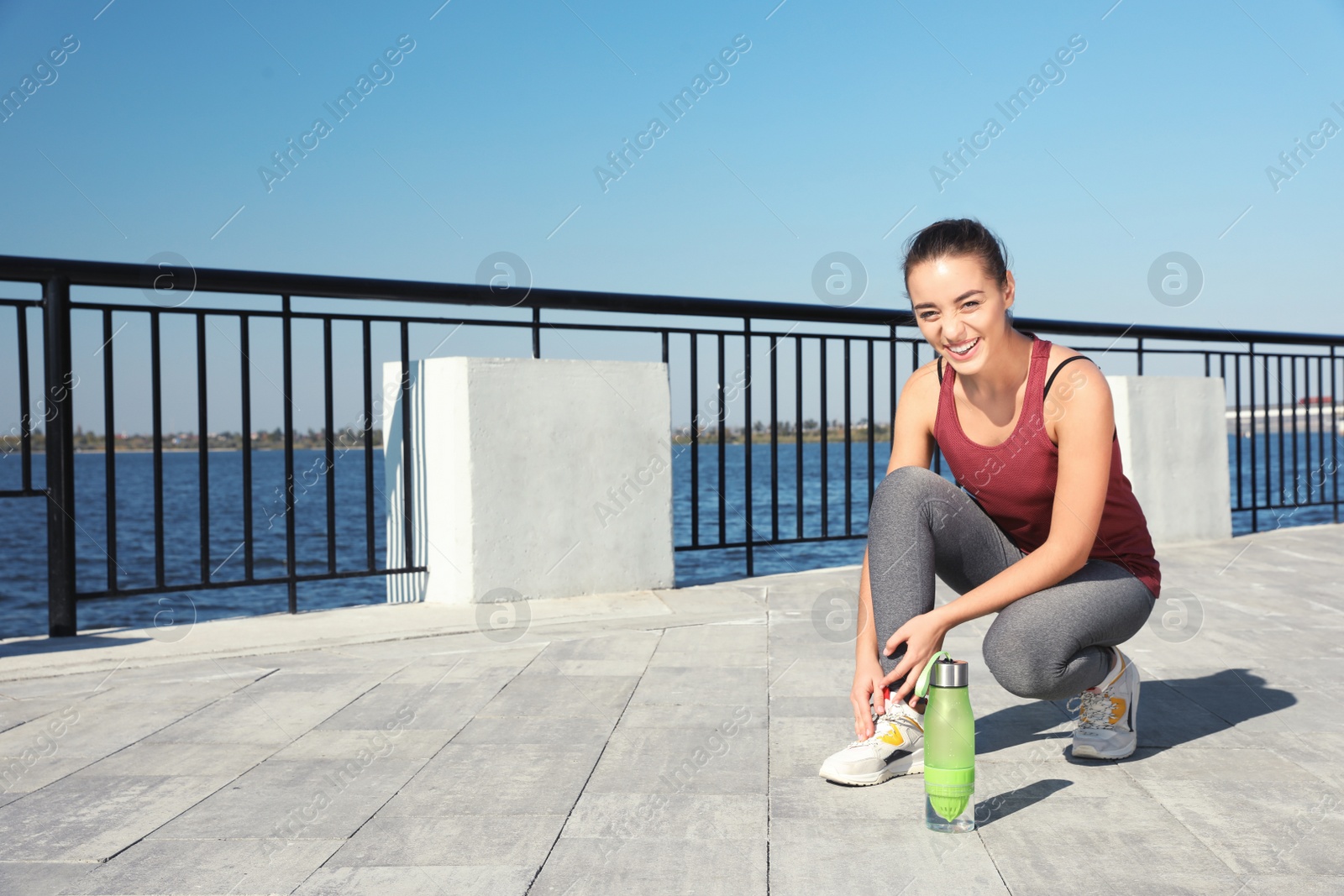 Photo of Young sporty woman tying shoelaces near bottle of water outdoors on sunny day