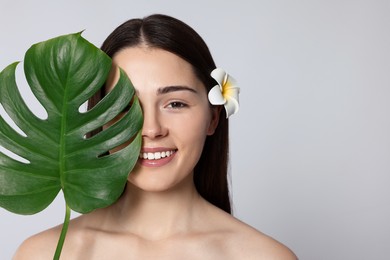 Photo of Young woman with plumeria flower in hair and leaf of monstera on light grey background, space for text. Spa treatment