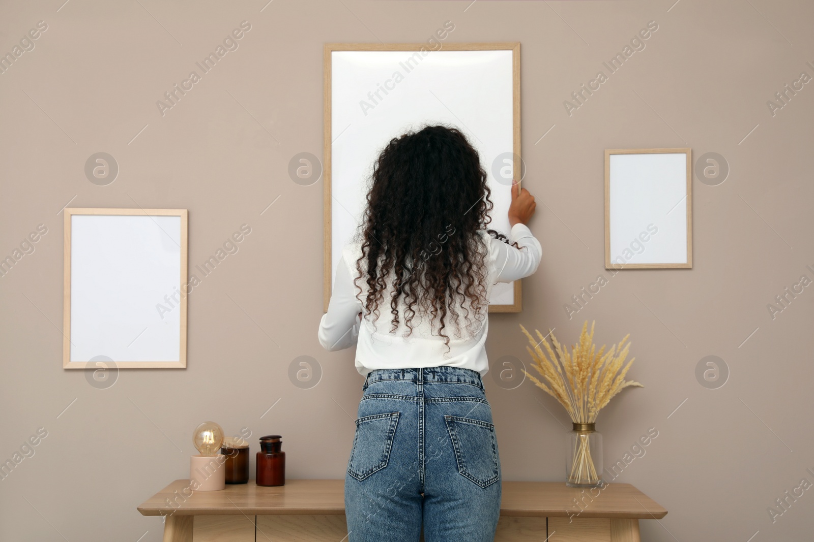Photo of Woman hanging empty frame on pale rose wall over table in room, back view
