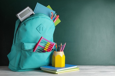 Stylish backpack with different school stationary on white wooden table at green chalkboard. Space for text