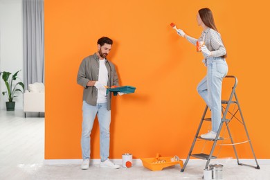 Photo of Woman painting orange wall and man holding container with roller indoors. Interior design
