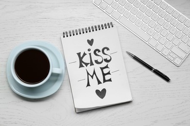 Photo of Notebook with phrase Kiss Me, keyboard, cup of drink and pen on white wooden table, flat lay