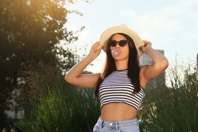 Beautiful woman in stylish sunglasses blowing gum outdoors on sunny day, space for text