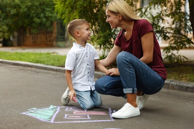 Photo of Nanny with cute little boy drawing house with chalks on asphalt