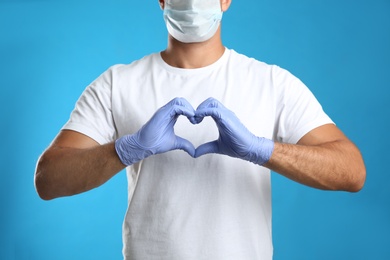 Man in protective face mask and medical gloves making heart with hands on blue background, closeup