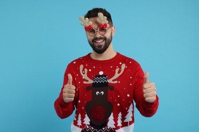 Photo of Happy young man in Christmas sweater and funny glasses showing thumbs up on light blue background