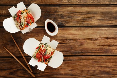 Photo of Boxes of vegetarian wok noodles with chopsticks and soy sauce on wooden table, flat lay. Space for text