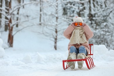 Photo of Cute little girl on sledge covering eyes with tangerines in snowy park, space for text. Winter vacation