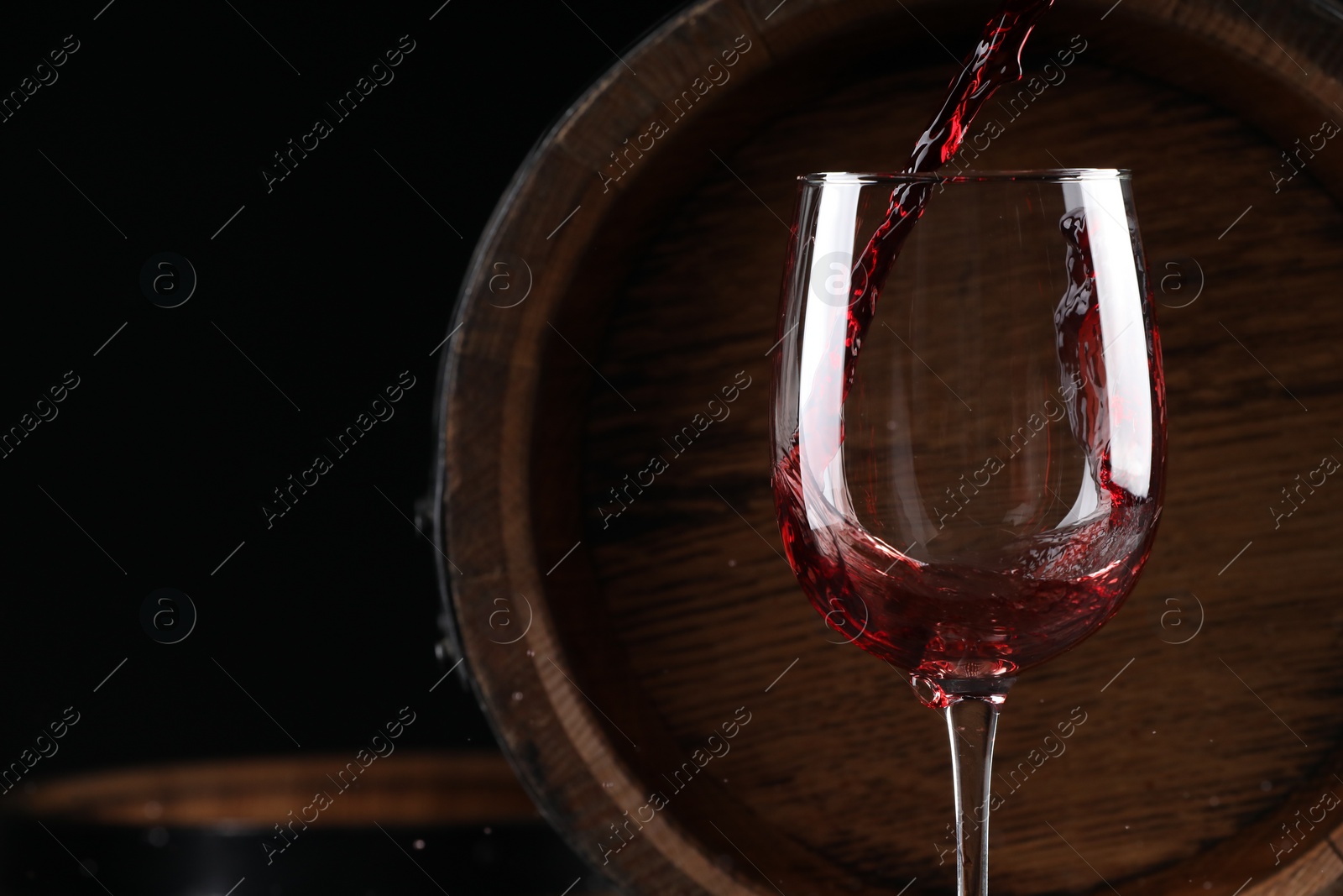 Photo of Pouring red wine into glass near wooden barrel against black background, closeup. Space for text