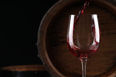 Photo of Pouring red wine into glass near wooden barrel against black background, closeup. Space for text