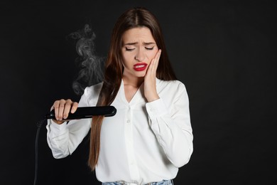 Upset young woman with flattening iron on black background. Hair damage
