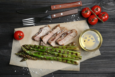 Photo of Tasty meat served with grilled asparagus on black wooden table, flat lay