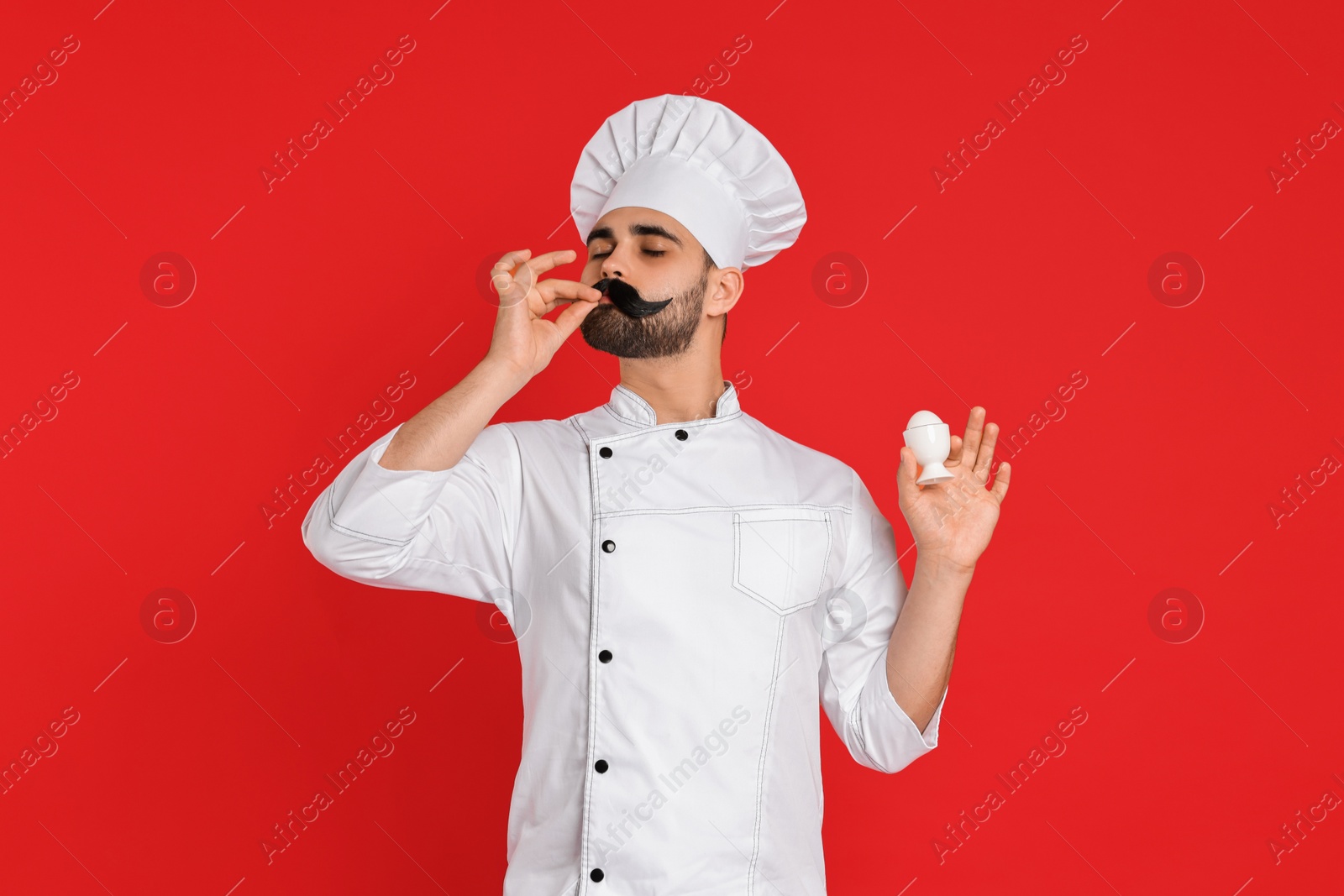 Photo of Professional chef with funny artificial moustache holding egg and showing perfect sign on red background