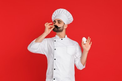 Photo of Professional chef with funny artificial moustache holding egg and showing perfect sign on red background