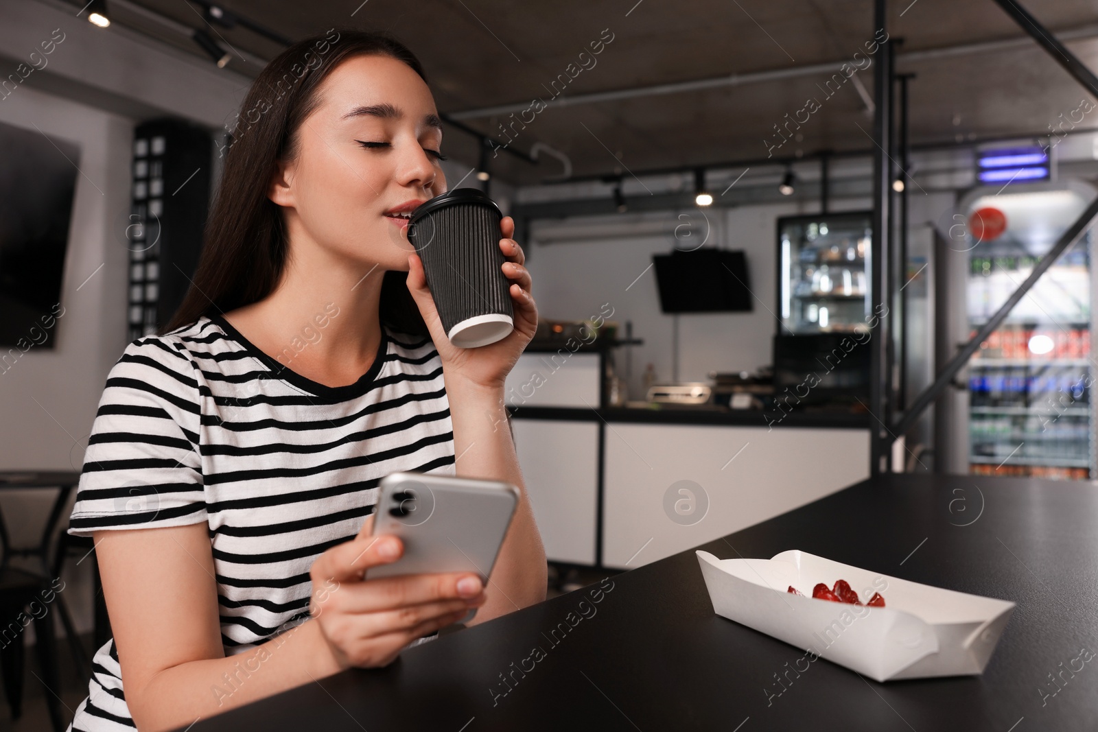 Photo of Happy young woman with paper cup of coffee and smartphone at table in hostel dining room