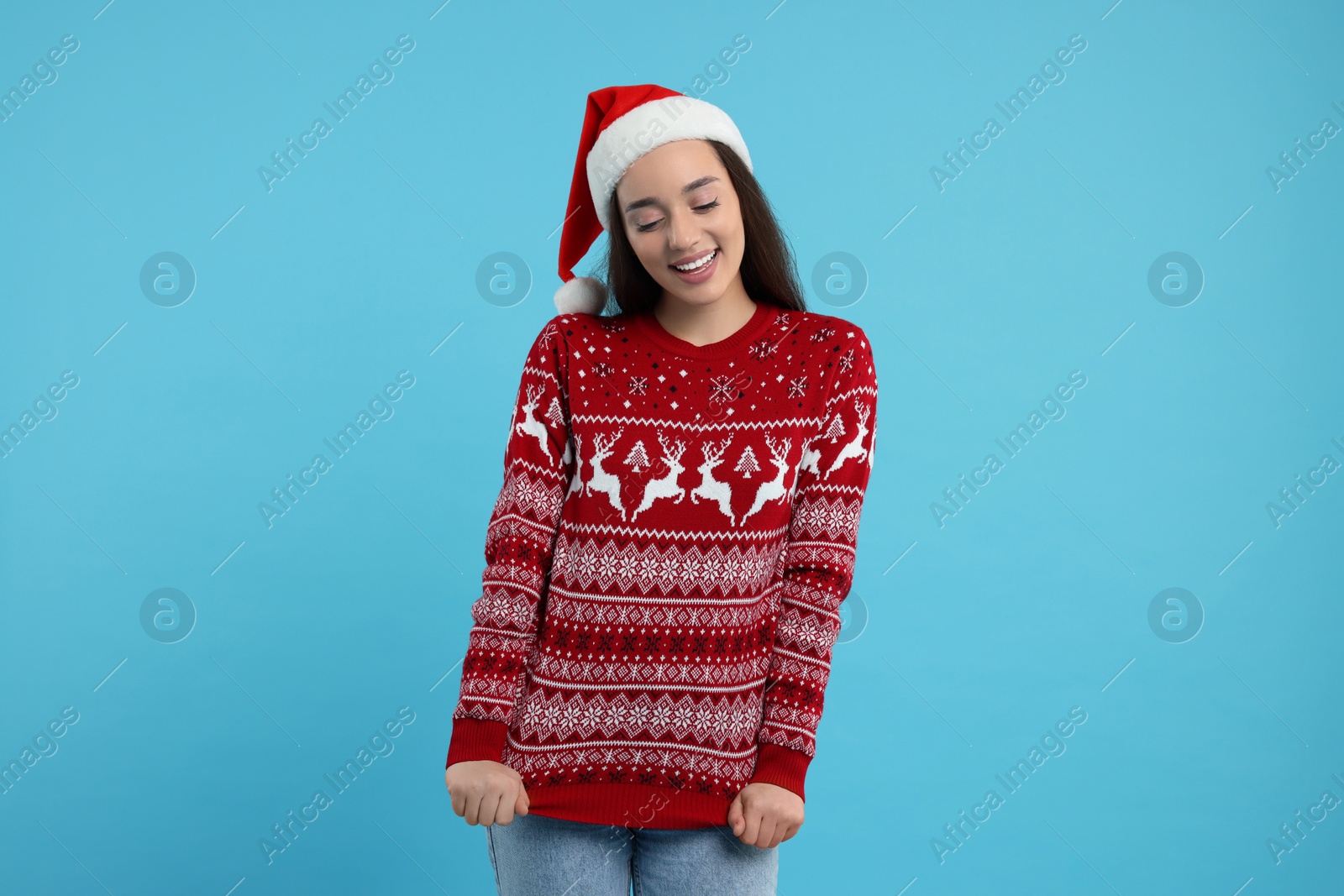 Photo of Happy young woman in Santa hat showing Christmas sweater on light blue background