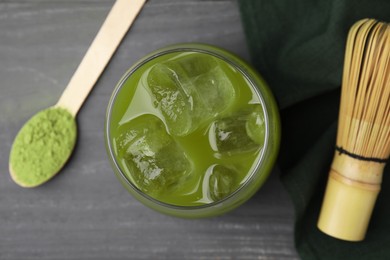 Photo of Glass of delicious iced green matcha tea, spoon with powder and bamboo whisk on grey wooden table, flat lay