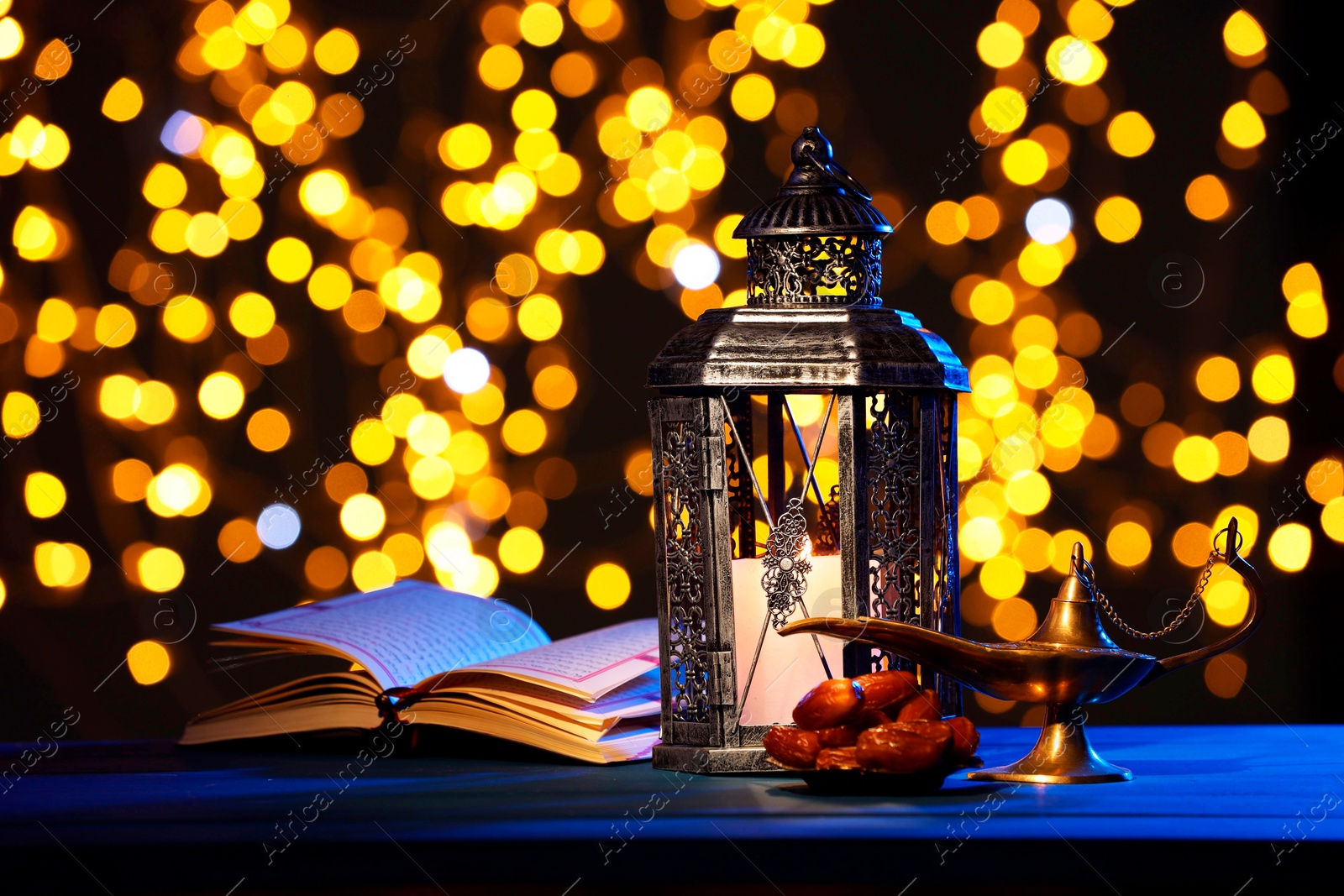 Photo of Arabic lantern, Quran, dates and Aladdin magic lamp on table against blurred lights at night