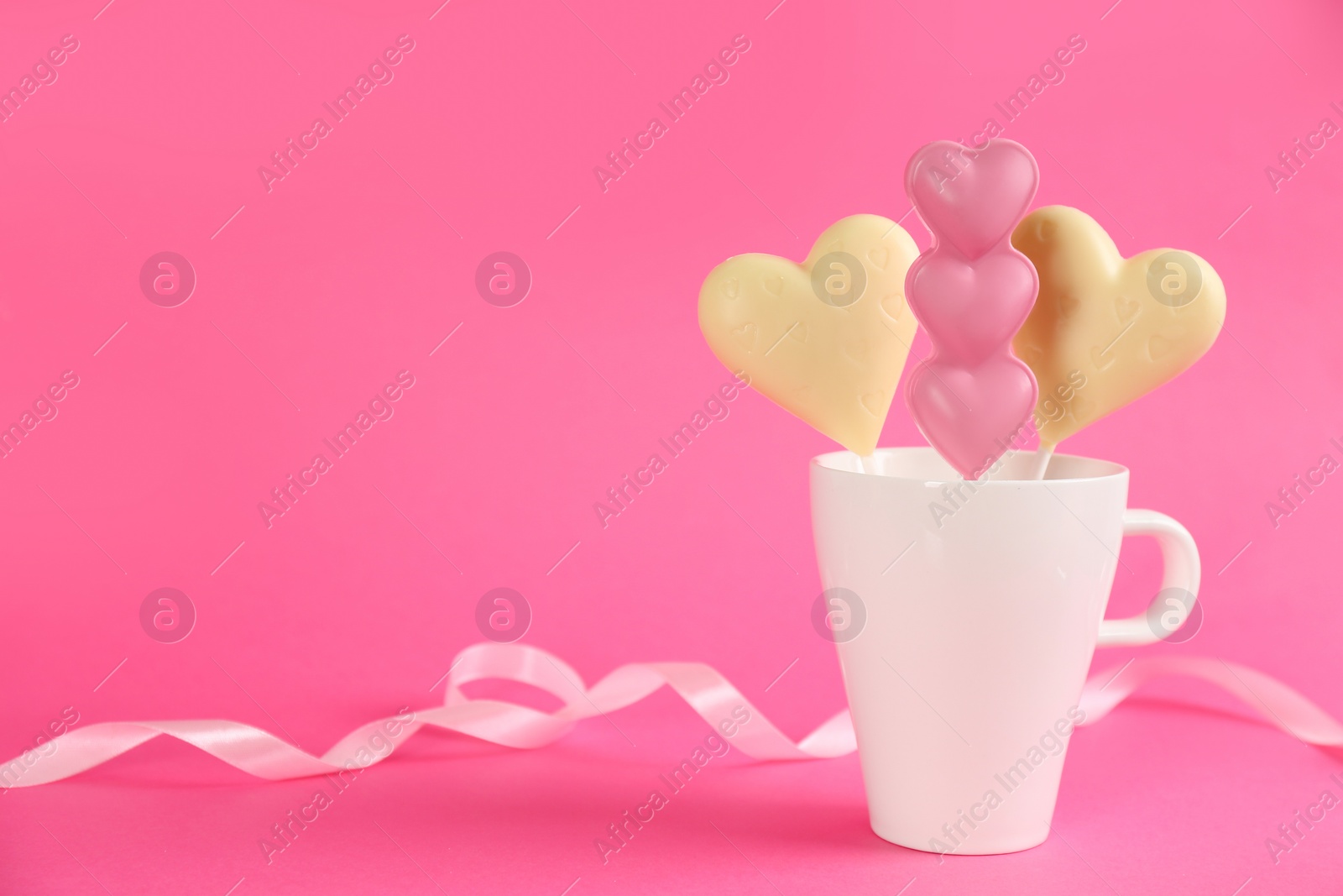 Photo of Heart shaped lollipops made of chocolate in cup and ribbon on pink background, space for text