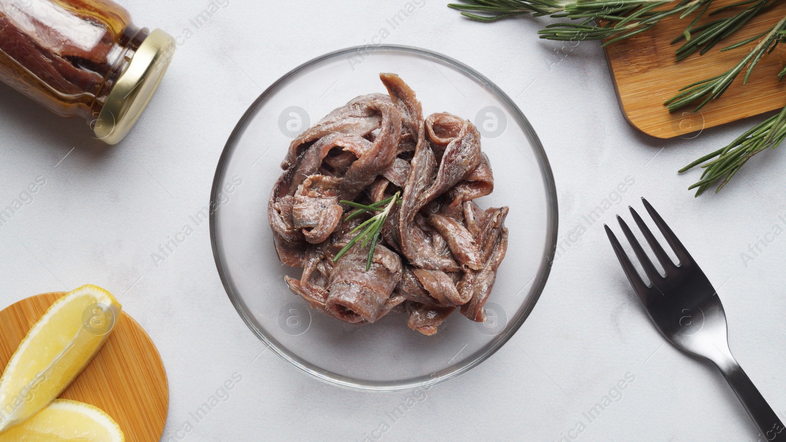 Photo of Canned anchovy fillets in glass bowl, rosemary and lemon wedges on white marble table, flat lay