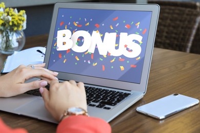 Image of Bonus gaining. Woman using laptop at wooden table indoors, closeup. Illustration of falling confetti and word on device screen