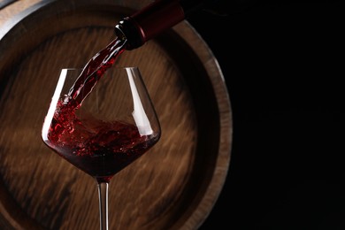 Photo of Pouring red wine from bottle into glass near wooden barrel against black background. Space for text