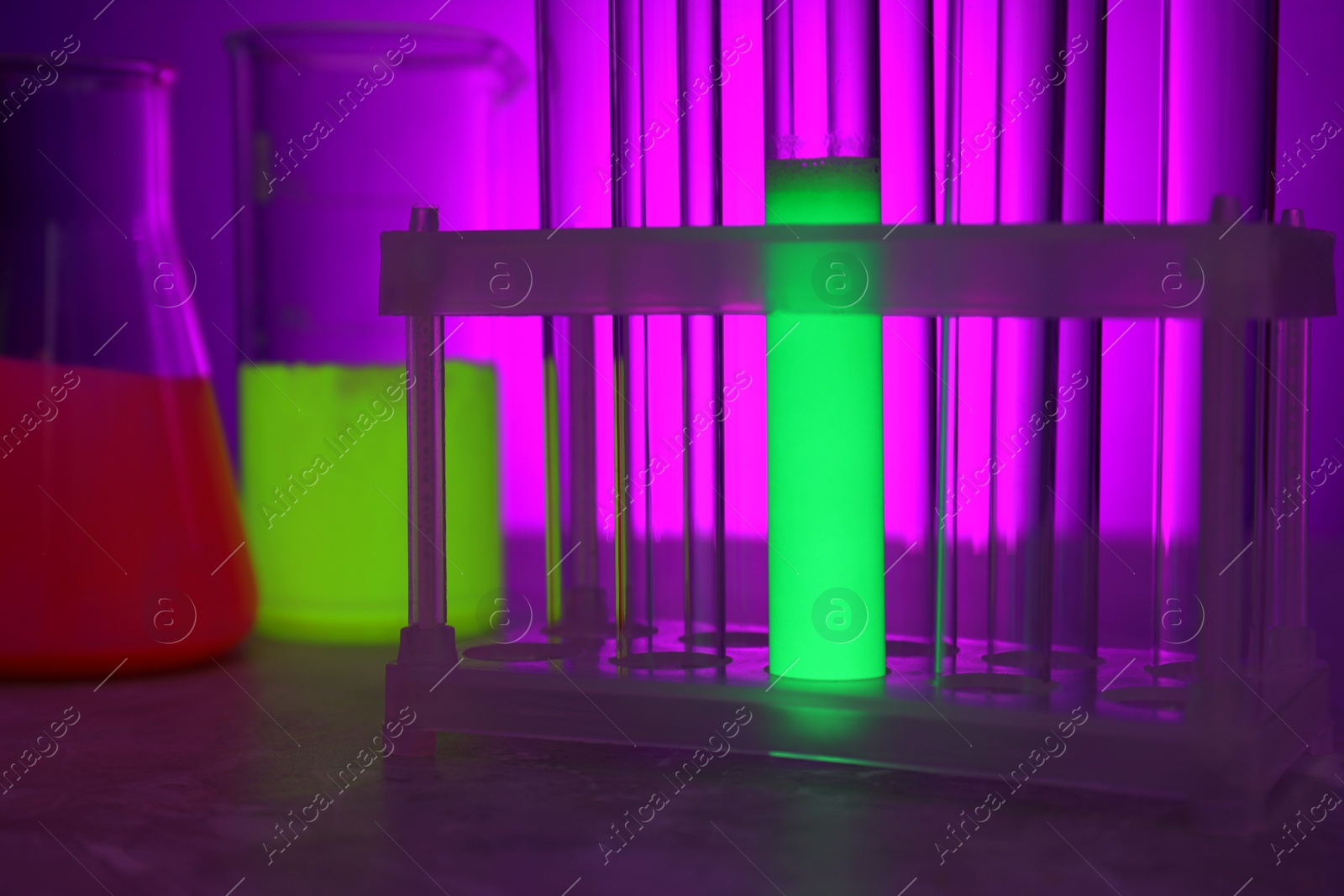 Photo of Laboratory glassware with luminous liquids on table against violet background, closeup