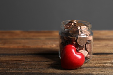 Red heart and donation jar with coins on wooden table against grey background. Space for text