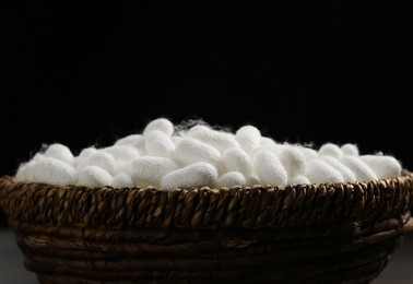 Photo of White silk cocoons in wicker bowl on black background, closeup