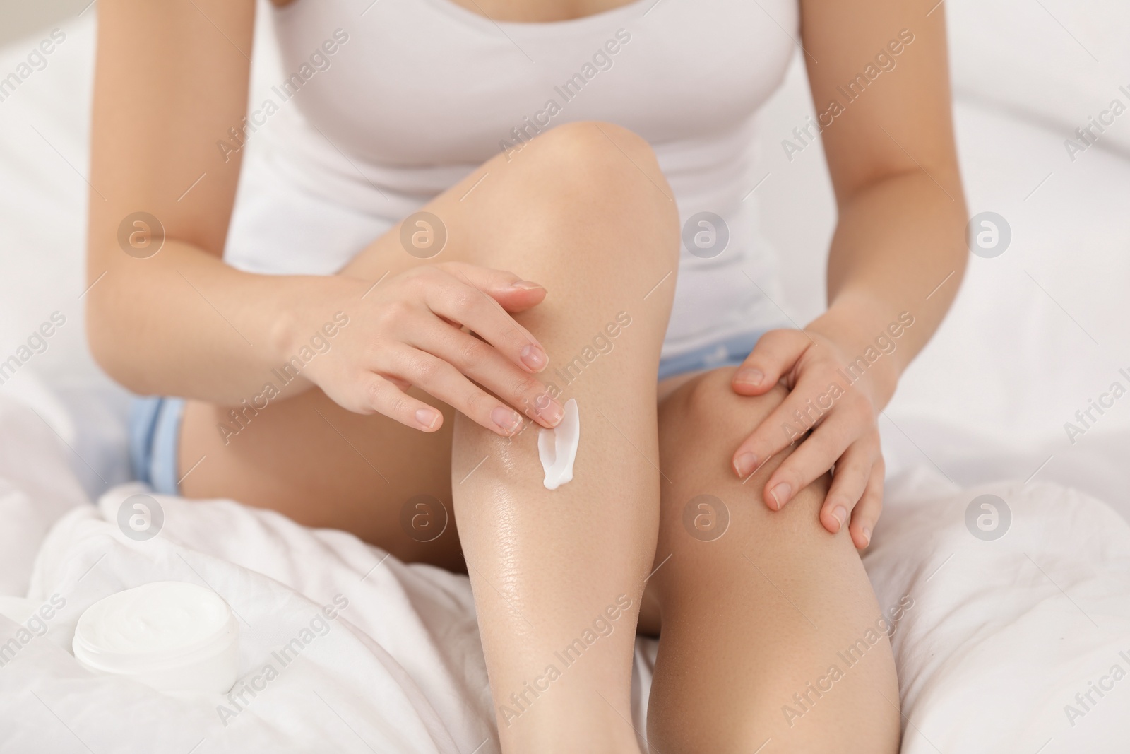 Photo of Young woman applying body cream onto leg on bed, closeup