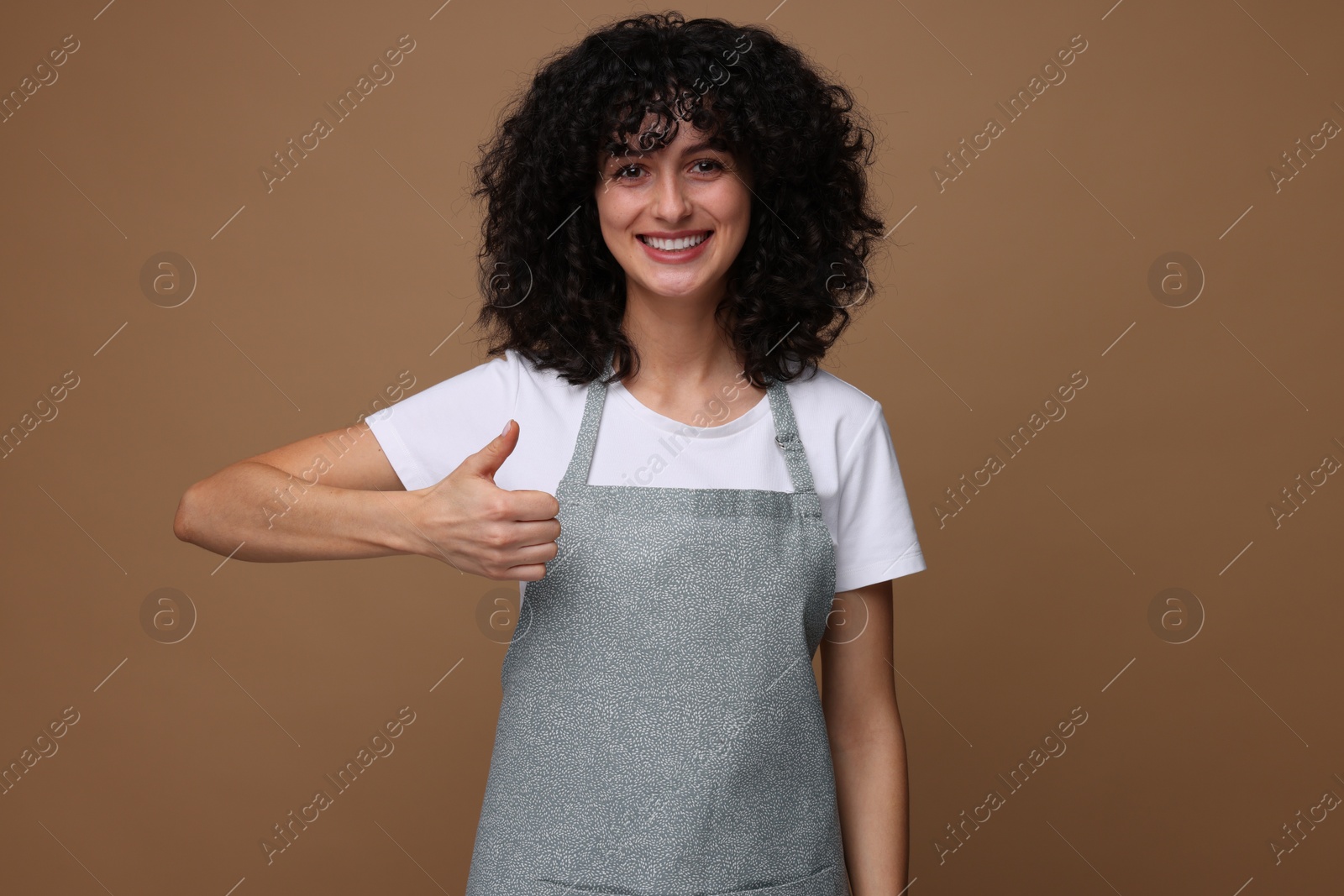 Photo of Happy woman wearing kitchen apron and showing thumbs up on brown background. Mockup for design