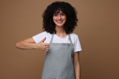 Photo of Happy woman wearing kitchen apron and showing thumbs up on brown background. Mockup for design