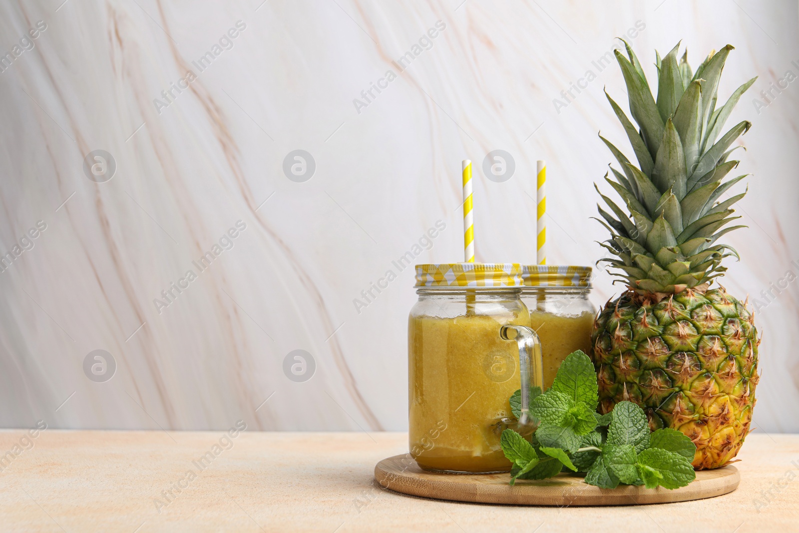 Photo of Tasty pineapple smoothie in mason jars, mint and fruit on beige table against marble background. Space for text