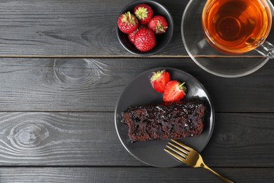 Photo of Piece of tasty chocolate sponge cake with strawberries and tea on black wooden table, flat lay. Space for text