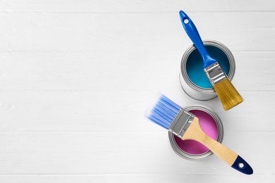 Cans of pink and light blue paints with brushes on white wooden table, flat lay. Space for text