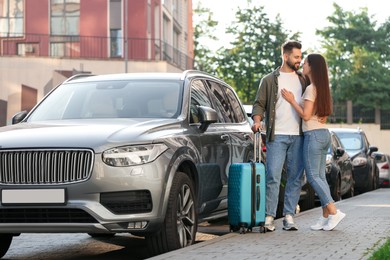 Photo of Long-distance relationship. Beautiful young couple with luggage hugging near car outdoors