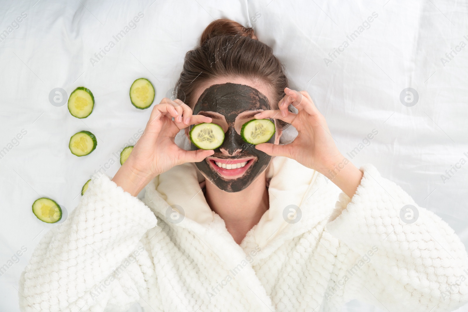 Photo of Young woman with facial mask lying and cucumber slices on bed, top view